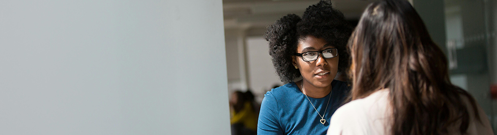 A counselor and her client sitting at a table, talking with eachother. The photo looks as if it was taken from around the corner of a wall.