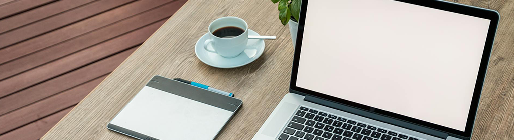 White-screened laptop sitting on a wooden table with a drawing pad, cup of tea, and a small plant.